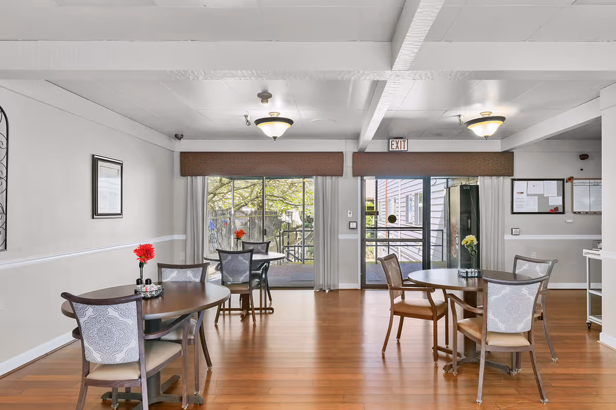 A bright dining area with wooden floors, several round and oval tables each surrounded by chairs with patterned backs. Each table has a small vase with flowers. Large sliding glass doors at the back lead to an outdoor area, and there is an exit sign above one door. The walls are light-colored with some framed artwork and bulletin boards.