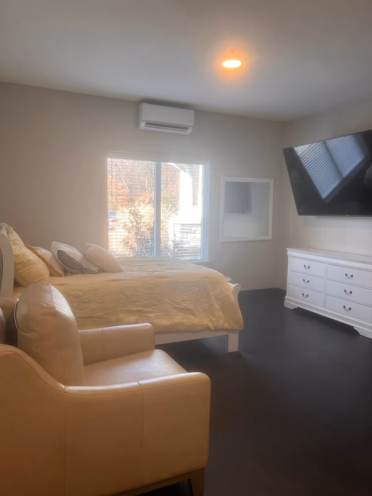 A bedroom with a large bed covered in a beige quilt and multiple pillows, a beige armchair in the foreground, a white dresser with drawers, a large flat-screen TV mounted on the wall, a window with blinds letting in natural light, and an air conditioning unit above the window.
