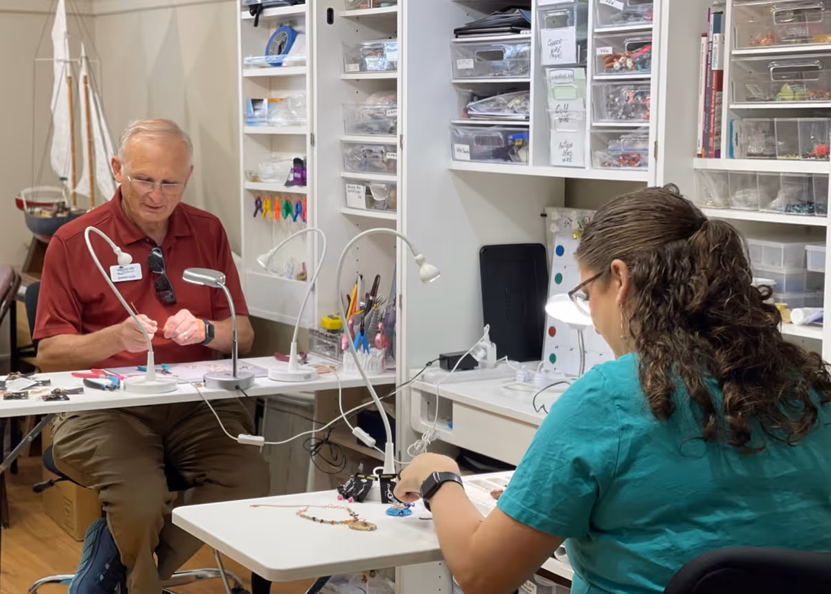 Two people sitting at separate tables in a well-lit craft room, working on jewelry making. The man on the left wears a red shirt and glasses, while the woman on the right wears a teal shirt and glasses. Shelves filled with organized craft supplies and tools are visible in the background.