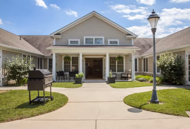 Exterior view of a single-story senior living facility building with beige siding, white trim, and a covered entrance supported by four white columns. There are two hanging plants on the porch, outdoor seating with chairs and small tables, a black grill on the left side, a lamppost on the right, and well-maintained green grass and shrubs surrounding the concrete walkway leading to the entrance.