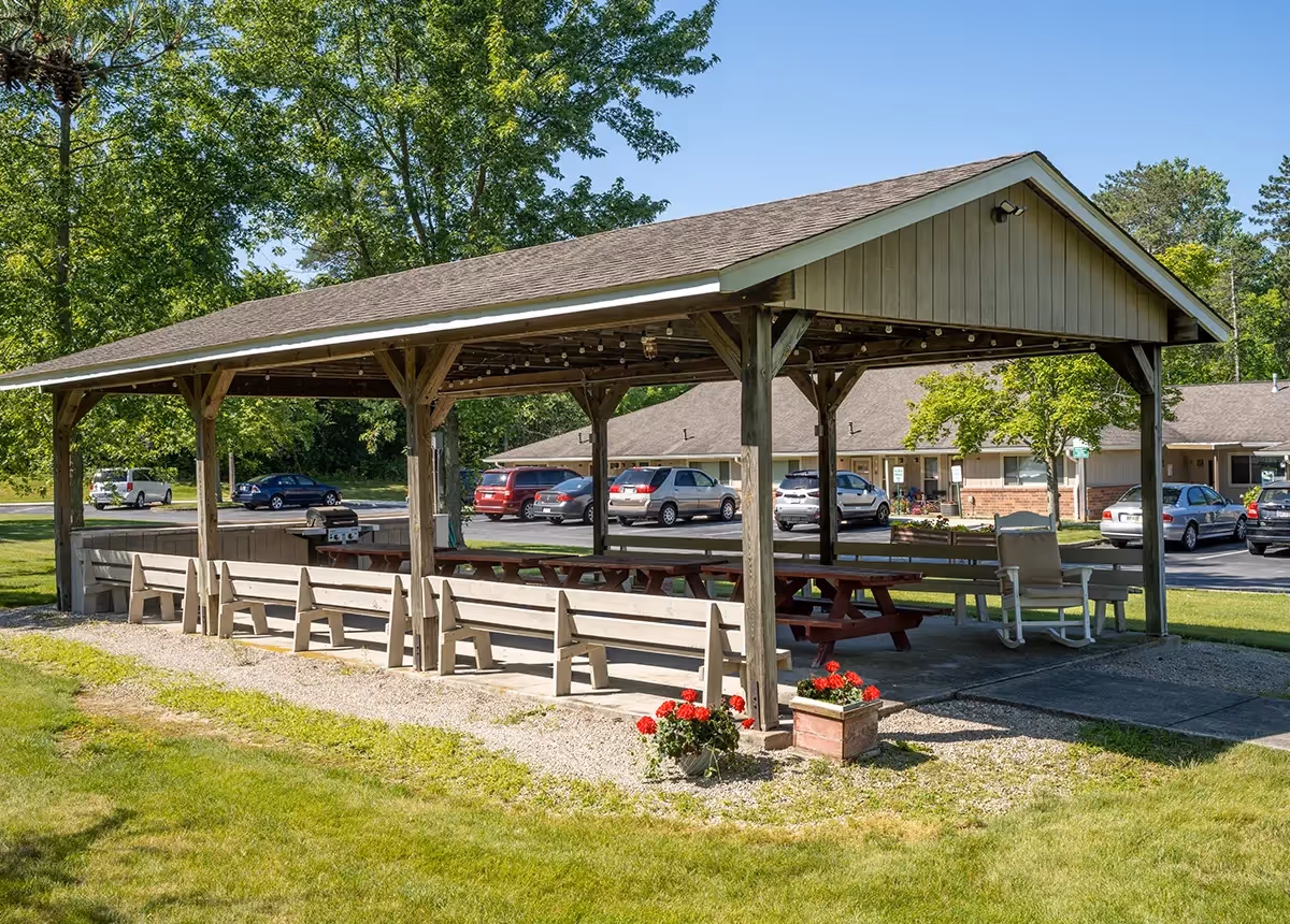 Outdoor covered pavilion with wooden benches and picnic tables, surrounded by grass and trees, with parked cars and residential buildings in the background under a clear blue sky.