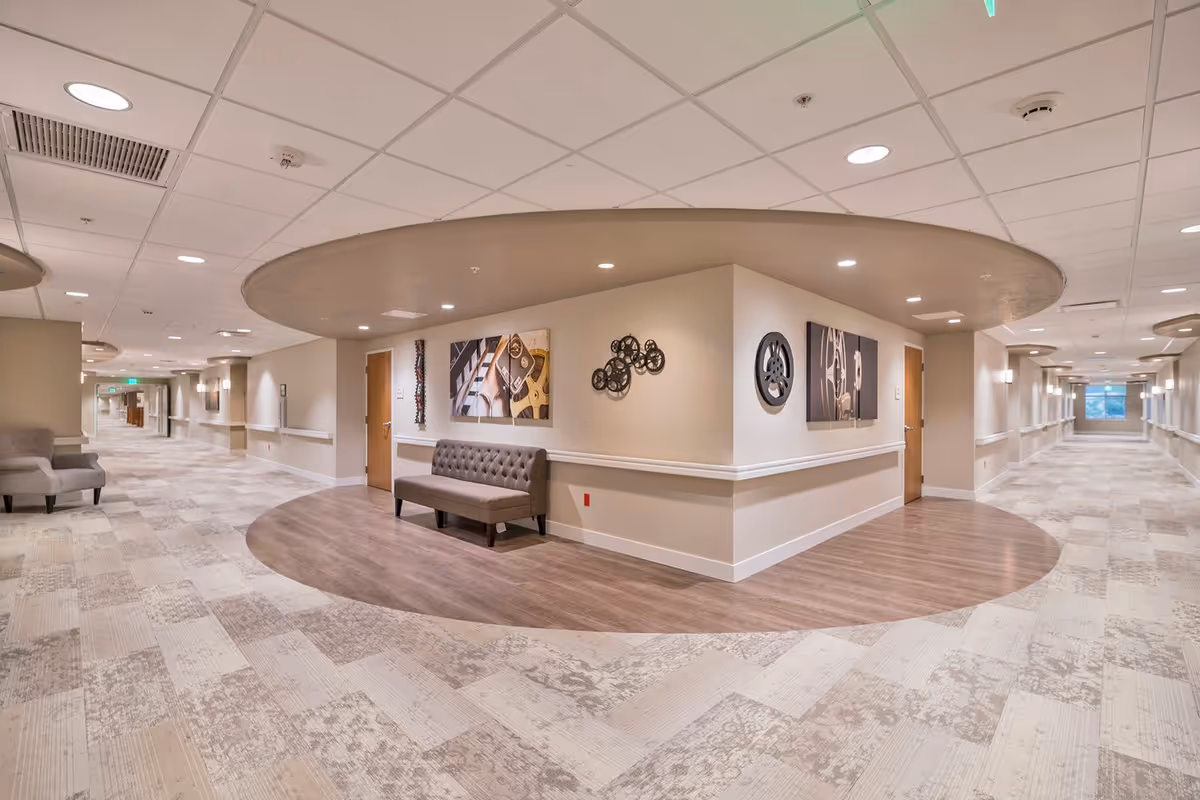A wide, well-lit hallway in a senior living facility with beige walls and patterned carpet flooring. The hallway features a circular recessed ceiling area with wood flooring underneath, housing a tufted bench and decorative wall art including abstract and gear-themed pieces. Doors line the hallway, and there are handrails along the walls for support.
