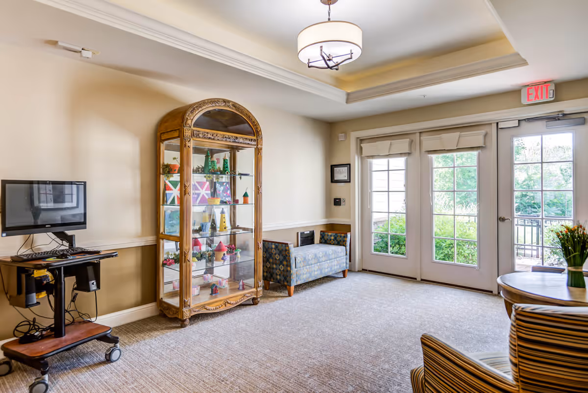 A bright and clean common area with beige walls and carpeted floor. There is a wooden display cabinet with glass doors showcasing colorful decorative items, a small patterned bench, a computer workstation on a rolling cart, and a round table with a vase of flowers. Large glass double doors lead outside to a green garden area, and an exit sign is visible above the doors.