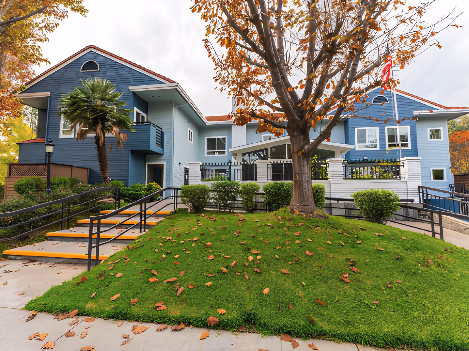 Exterior view of a two-story blue and white building with a red roof, surrounded by green grass, bushes, and trees with autumn leaves. There are stairs and ramps with black railings leading up to the entrance, and a flagpole with a flag is visible near the building.
