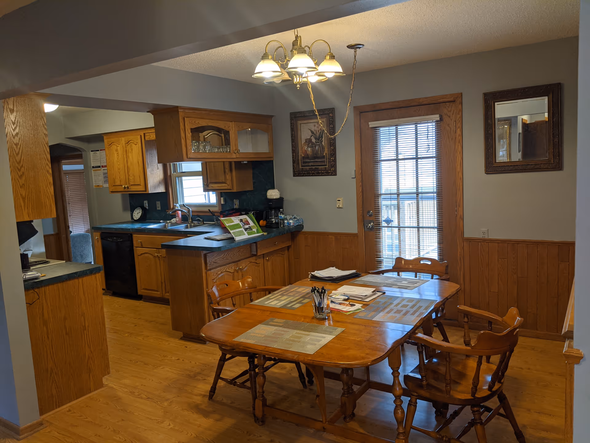 A cozy kitchen and dining area with wooden cabinets, a black countertop, and a wooden dining table with four chairs. The table has placemats, papers, and a container with pens. A glass door with blinds is visible in the background, along with framed artwork and a mirror on the walls. The floor is wooden, and a chandelier-style light fixture hangs above the dining table.