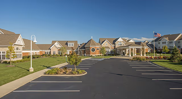 Exterior front view of a large senior living facility with a paved parking lot, landscaped lawns, and an American flag under a clear blue sky.