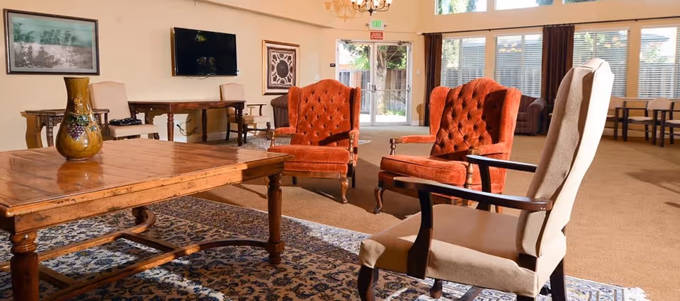 A spacious living room area with a wooden coffee table, two orange upholstered armchairs, and a beige armchair. The room has large windows with blinds, a patterned rug, a wall-mounted TV, and framed artwork on the walls. There is a door leading outside and additional seating in the background.