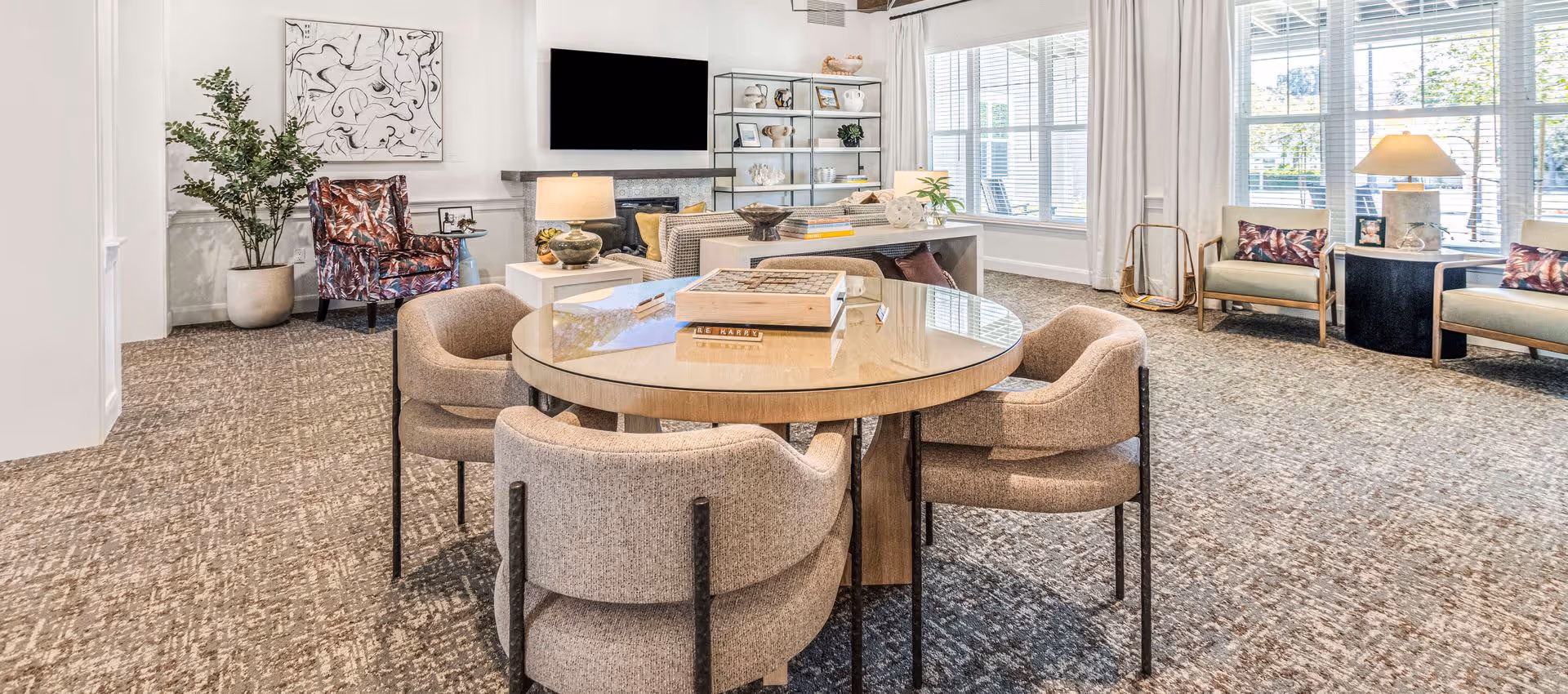 A bright and spacious living room with a round table surrounded by four upholstered chairs in the foreground. The room features a large window with white blinds, two armchairs with patterned pillows, a sofa, a side table with a lamp, a flat-screen TV mounted above a fireplace, and a shelving unit with decorative items. There is also a large potted plant and abstract wall art on the left side.