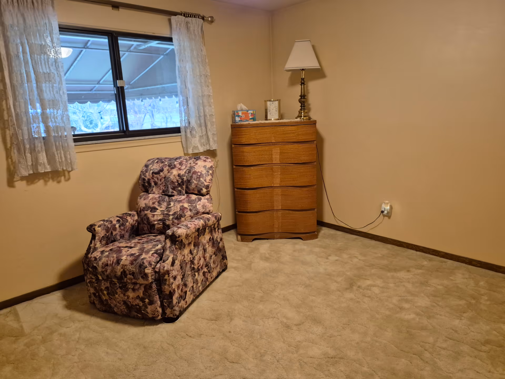 A small room with beige walls and carpet, featuring a floral patterned recliner chair near a window with lace curtains. In the corner, there is a wooden chest of drawers with a brass lamp, a clock, and a box of tissues on top. An electrical outlet with a plugged-in cord is visible on the wall.