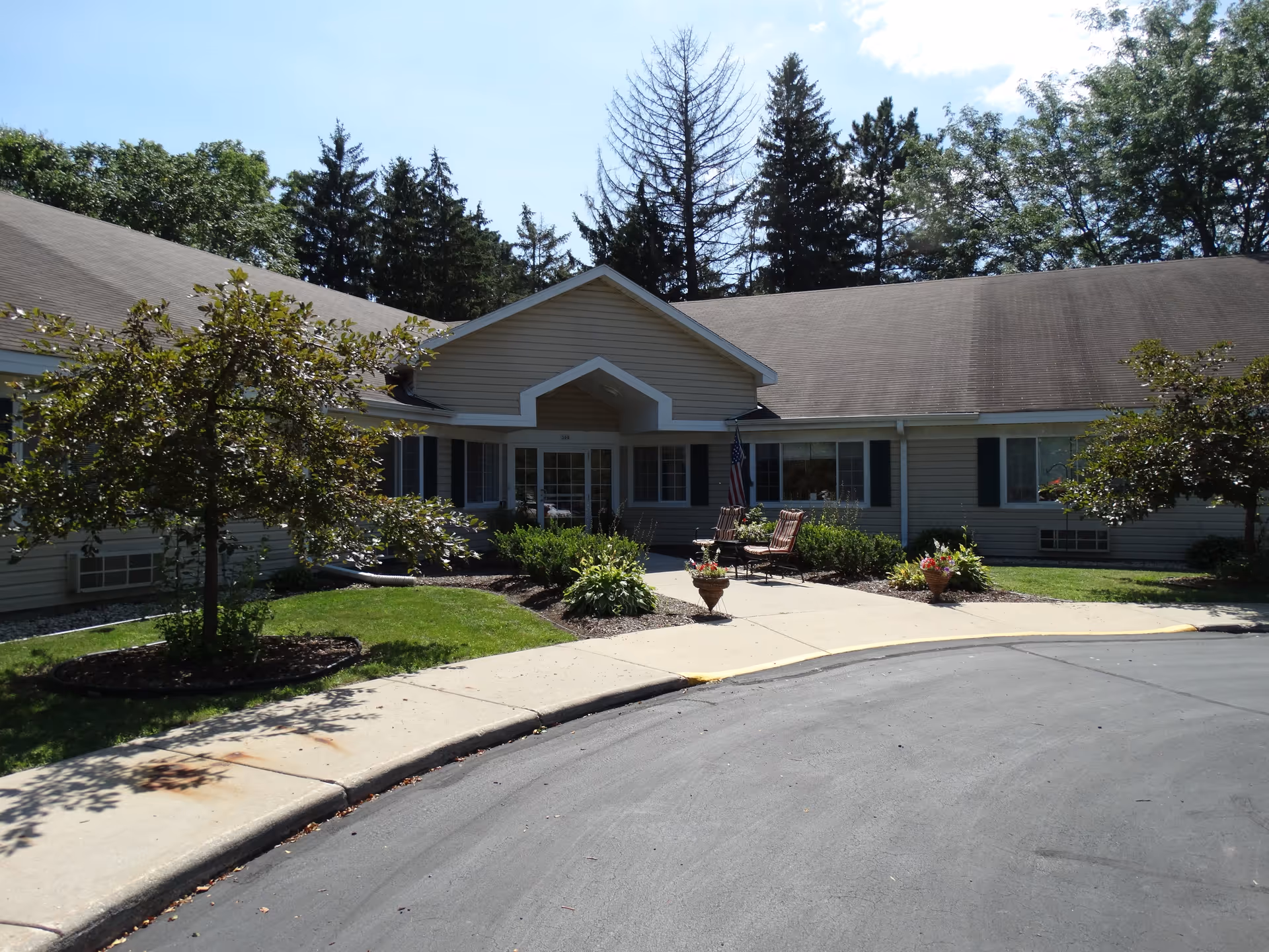 Exterior view of a single-story building with beige siding and a peaked entrance. There are small trees and landscaped bushes around the entrance, with a paved driveway and sidewalk leading up to the door. Two chairs and a small table are placed near the entrance, and tall trees are visible in the background under a clear sky.