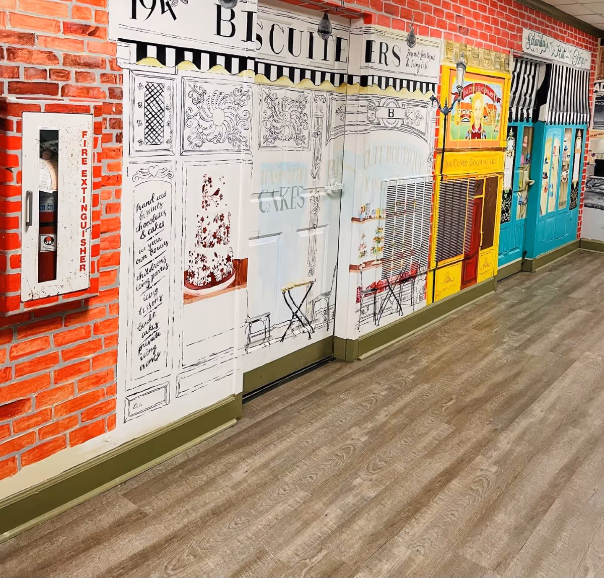 A hallway with walls painted to resemble storefronts of various shops, including a biscuit shop, a bakery, and other colorful storefronts. The floor is wood-patterned, and there is a fire extinguisher mounted on the brick-patterned wall on the left side.