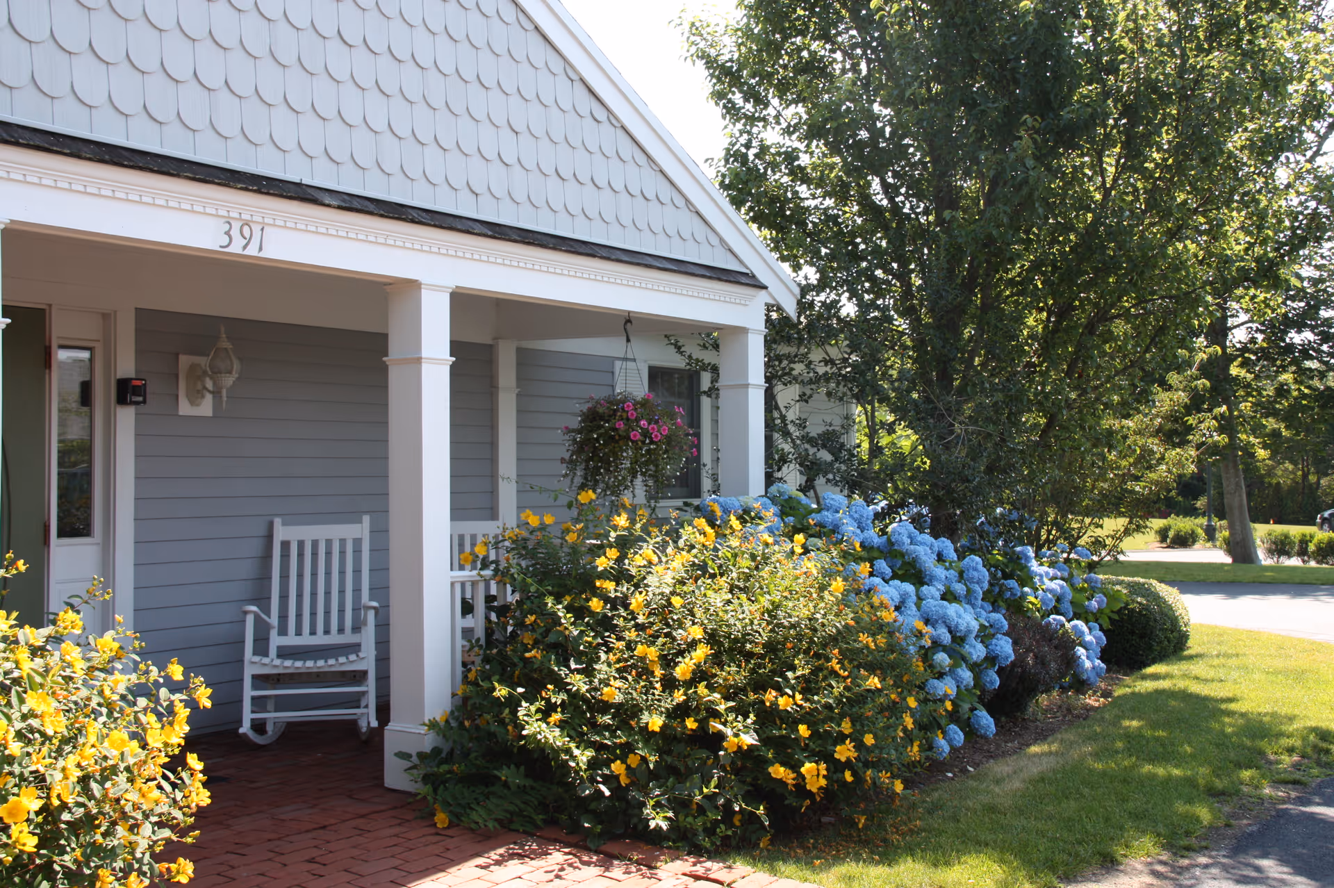 Front porch of a building with gray siding and white trim, featuring two white rocking chairs. The porch is surrounded by lush bushes with yellow and blue flowers, and there is a hanging basket with pink flowers. The building number 391 is visible above the porch.