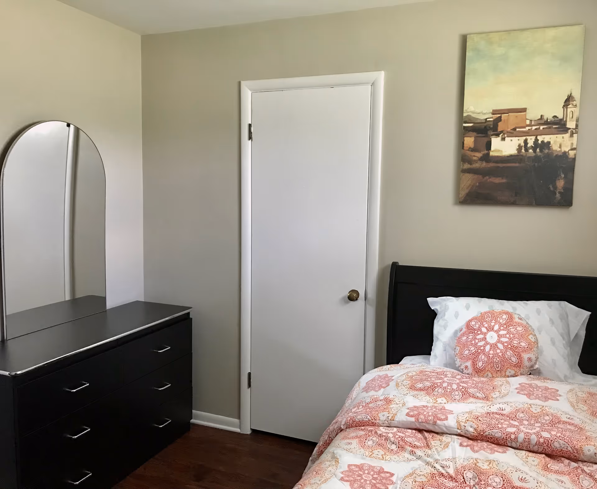 A tidy bedroom with a bed dressed in patterned bedding, a black dresser with a mirror, a closed white door, and wall art.
