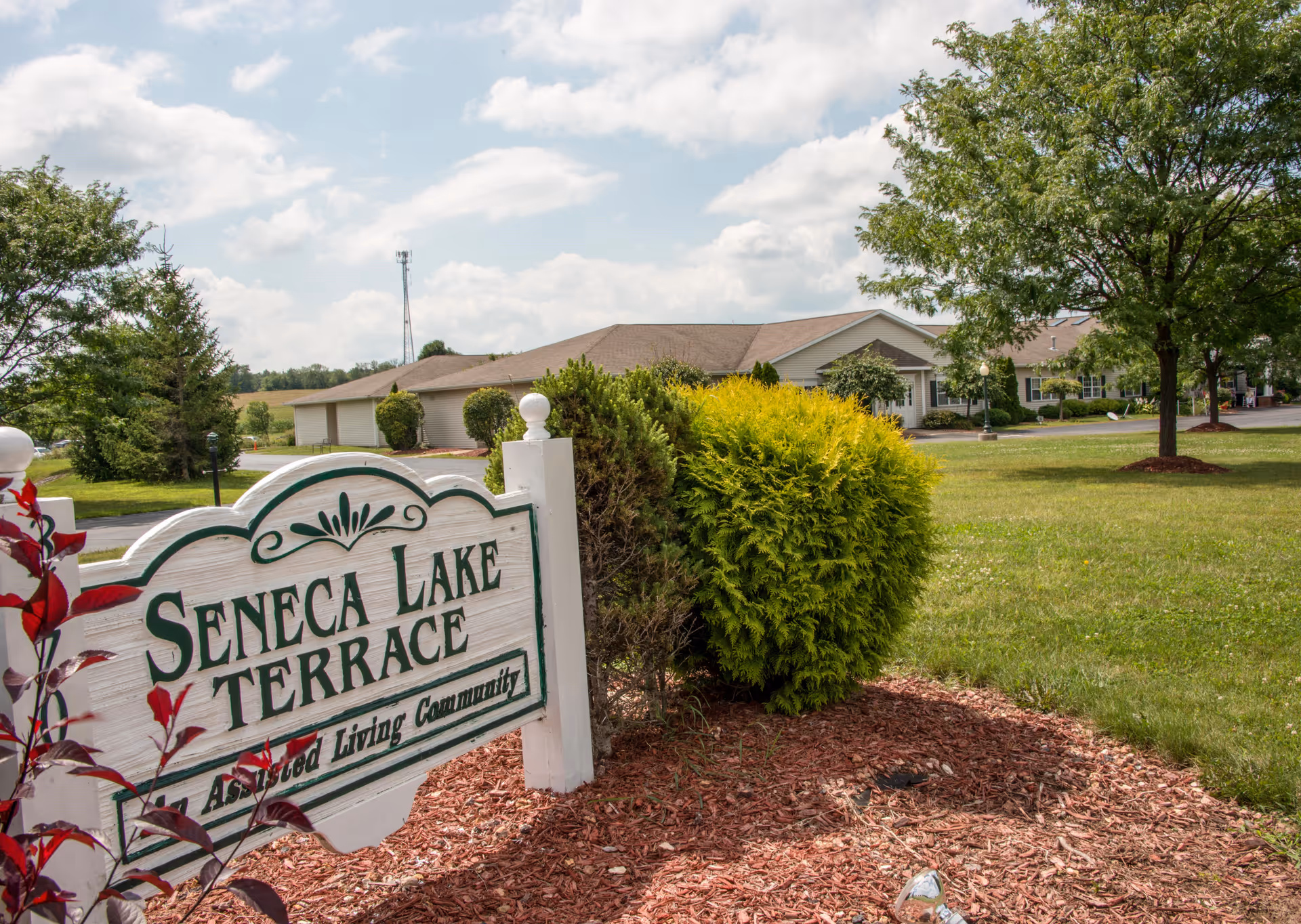Outdoor view of Seneca Lake Terrace assisted living community sign with a building and green lawn in the background under a partly cloudy sky.