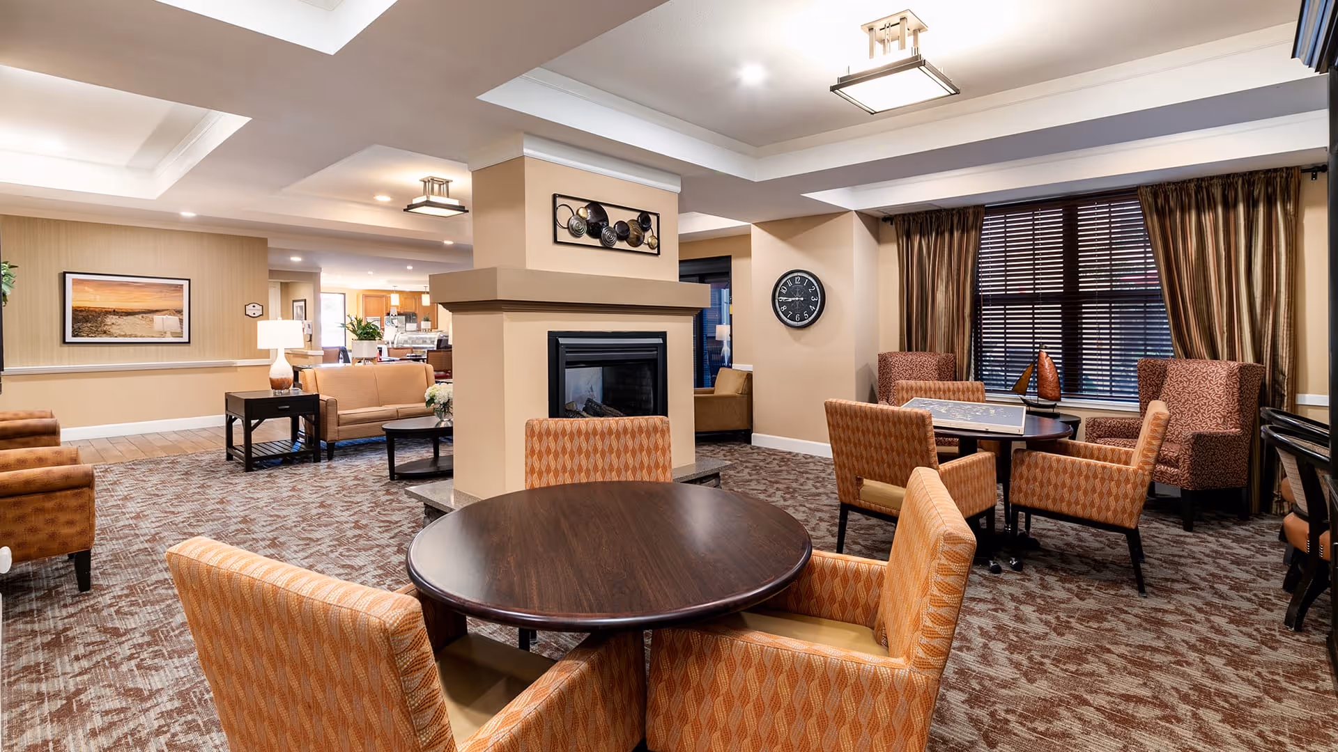 A spacious and well-lit living room area in a senior living facility with multiple seating arrangements including round tables with orange patterned chairs, armchairs, and sofas. The room features a central fireplace, a wall clock, large windows with brown curtains, and decorative wall art. The flooring is carpeted with a patterned design, and the ceiling has recessed lighting and modern light fixtures.