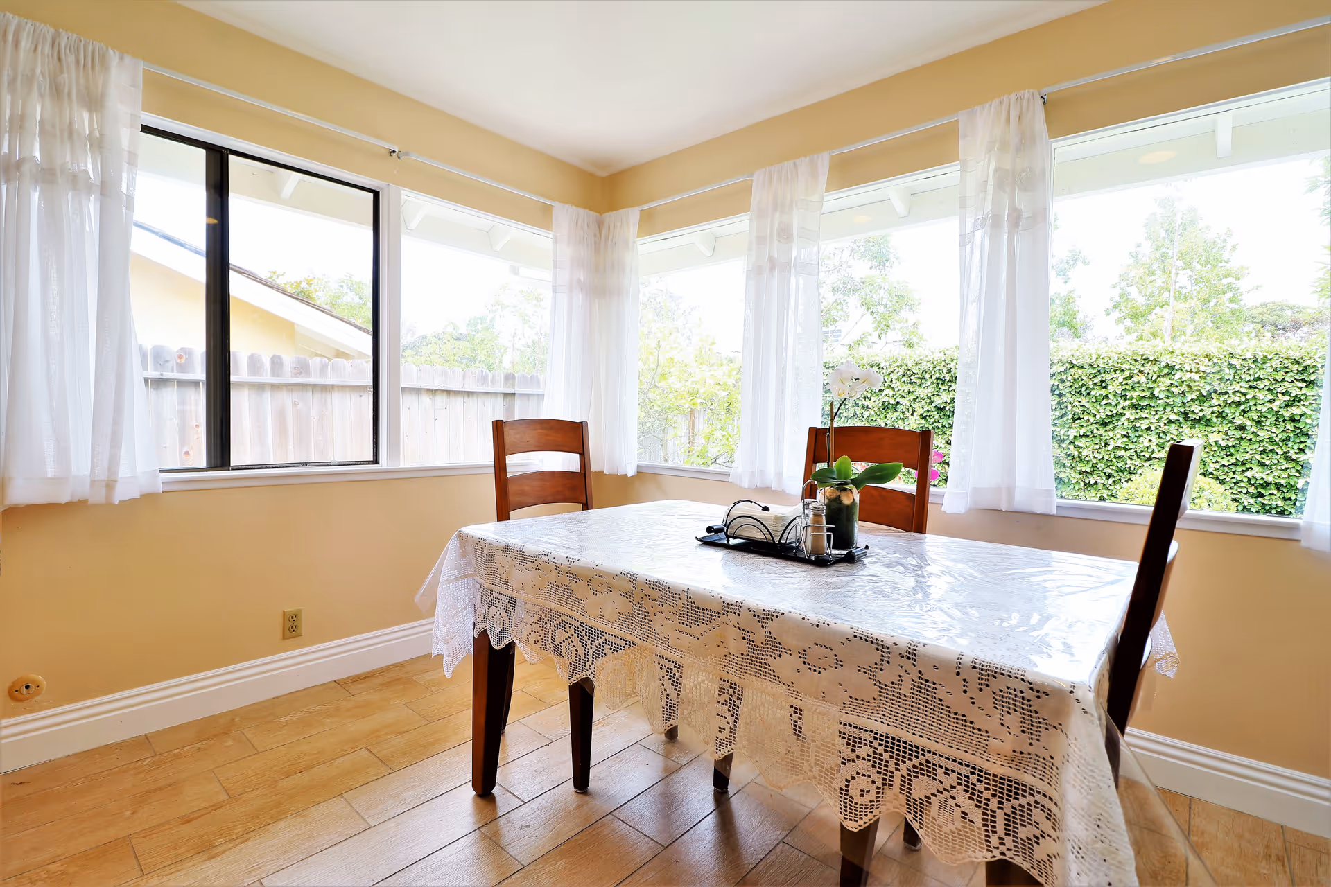 A bright dining area with large windows covered by sheer white curtains. There is a wooden table with a lace tablecloth and three wooden chairs around it. On the table, there is a small plant and a tray with salt and pepper shakers. Outside the windows, greenery and a wooden fence are visible.