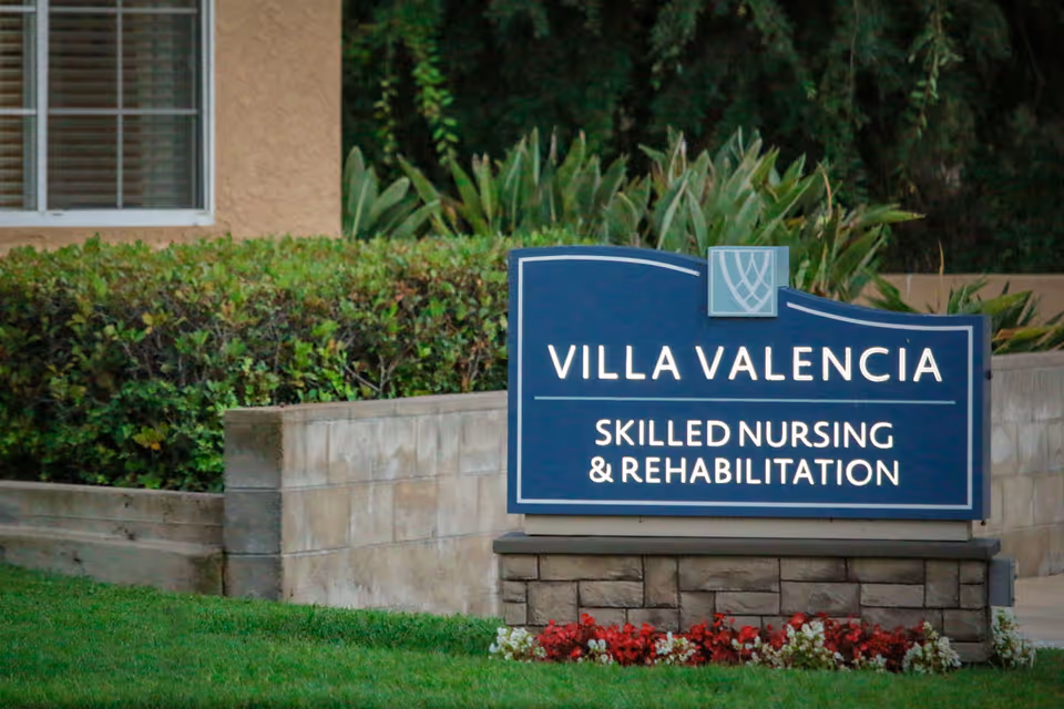 Outdoor sign for Villa Valencia Skilled Nursing & Rehabilitation facility surrounded by greenery and flowers.