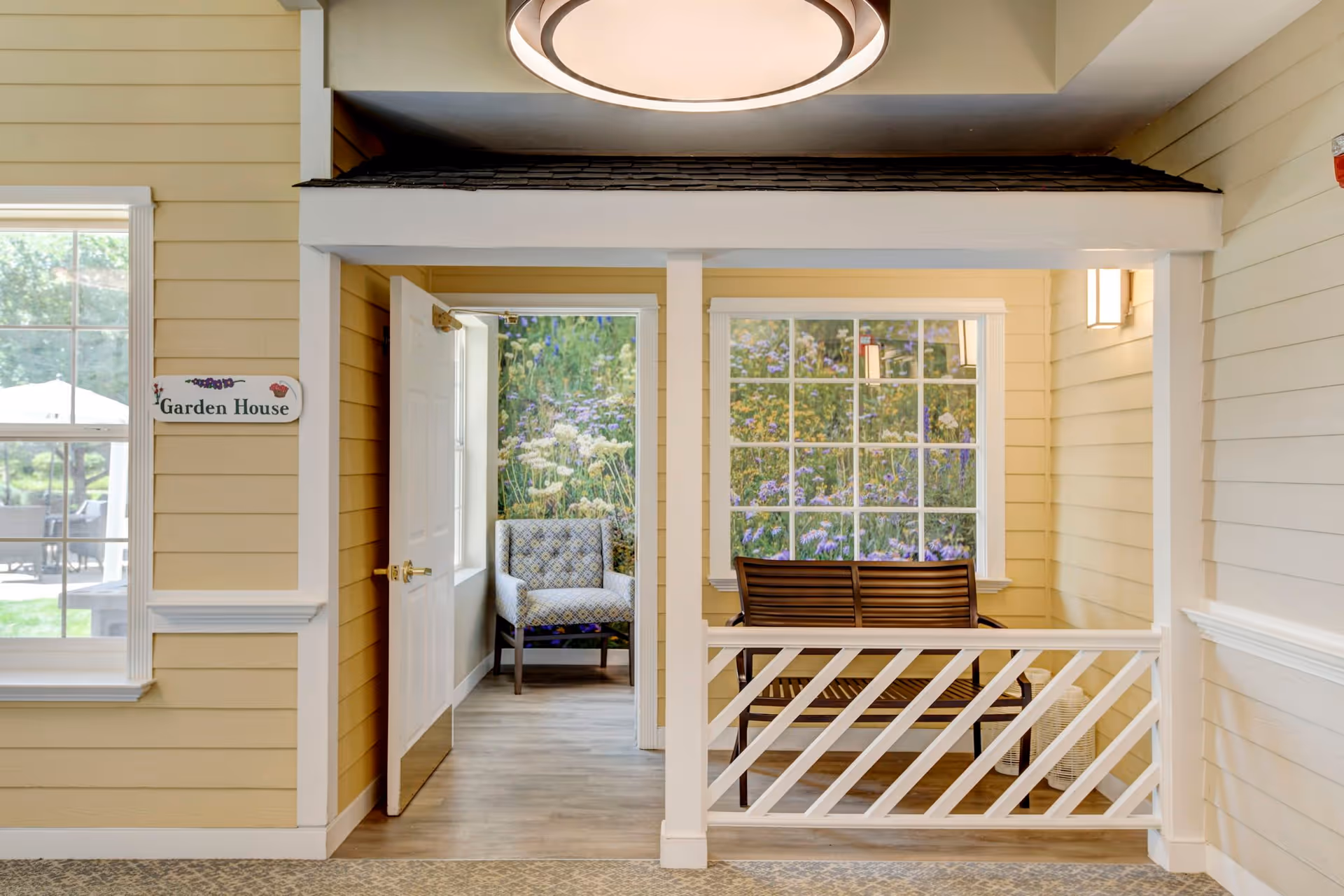 Indoor area designed to resemble a small garden house with beige walls and white trim. There is a white door open to a small room with a patterned armchair and a large window showing a floral mural. Outside the room is a wooden bench with a white railing in front and another large window with a floral mural. A sign on the wall reads 'Garden House'.