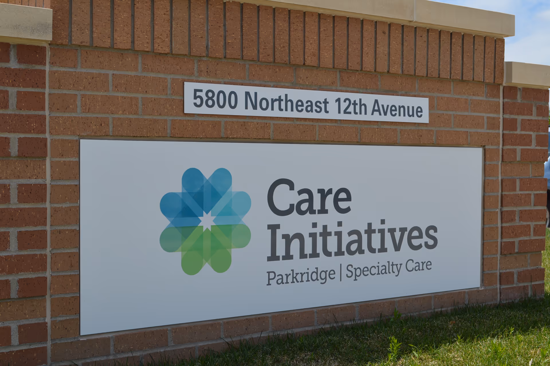 A brick signpost with a white sign displaying the address '5800 Northeast 12th Avenue' and a logo with the text 'Care Initiatives Parkridge | Specialty Care' in front of a grassy area under a clear sky.
