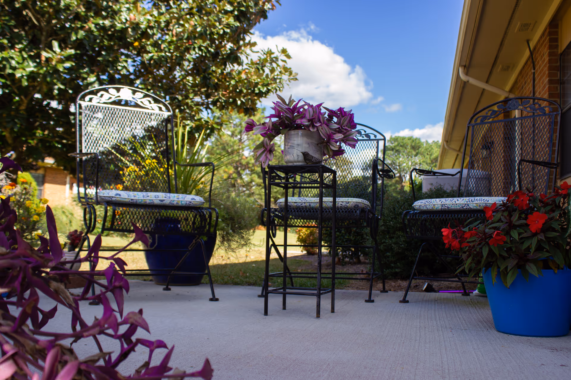 Patio with wrought-iron chairs, a small table holding a potted plant, and colorful planters under a blue sky.