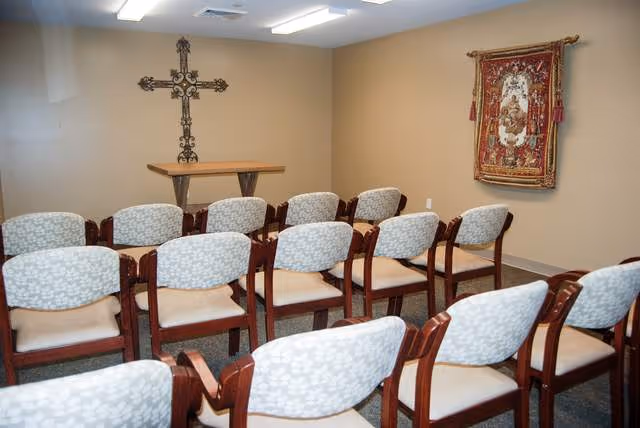 Small chapel room with rows of wooden chairs with light patterned cushions facing a wooden altar table. A decorative cross hangs on the wall behind the altar, and a tapestry with religious imagery is displayed on the adjacent wall.