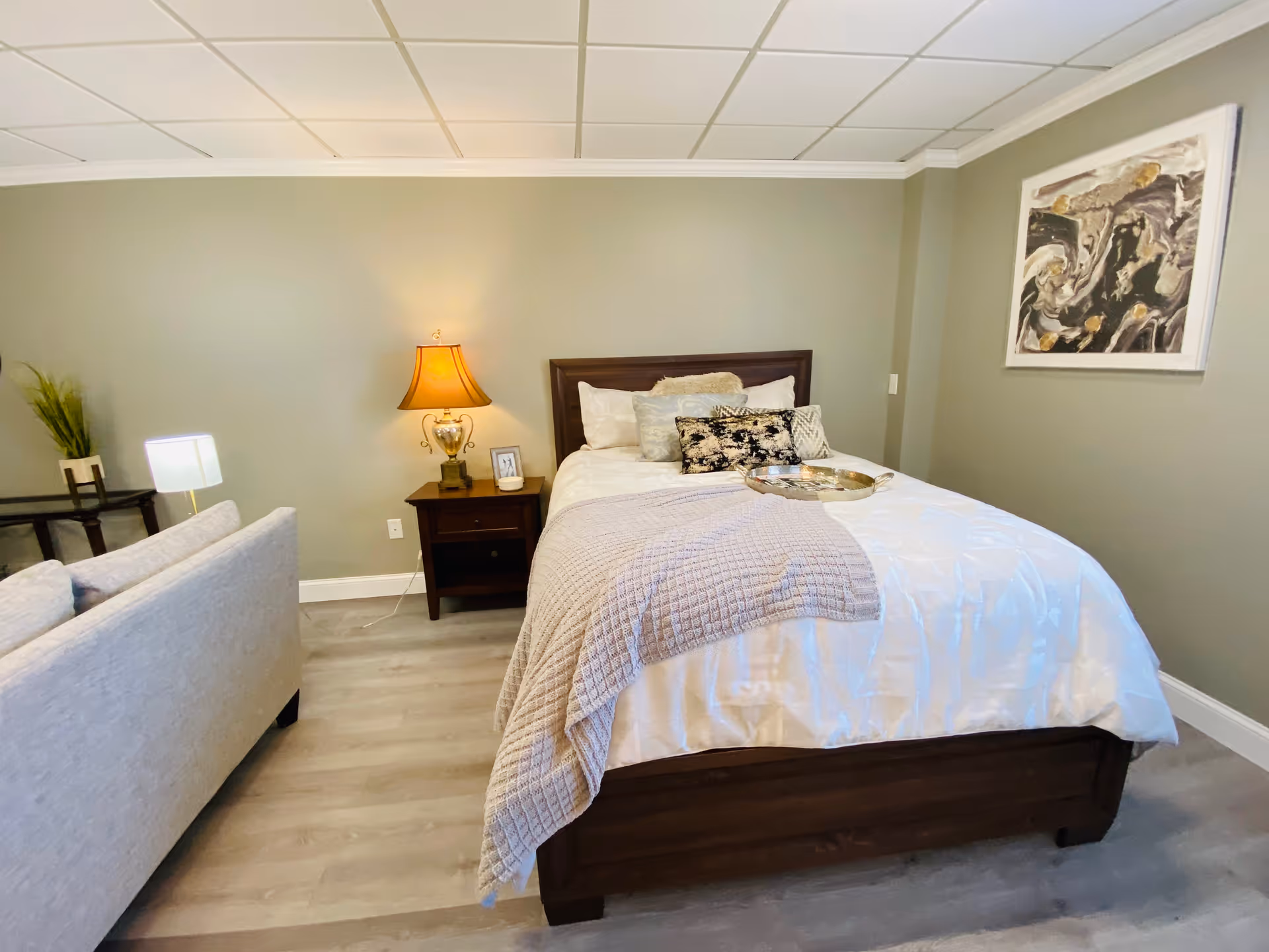 A cozy bedroom with a neatly made bed featuring white bedding and decorative pillows. A beige throw blanket is draped over the bed. There is a wooden nightstand with a lamp and a small framed photo. A light gray sofa is positioned at the foot of the bed. The walls are painted a soft gray-green color, and a modern abstract painting hangs on the wall to the right. The floor is light wood, and the ceiling has a grid pattern with recessed lighting.