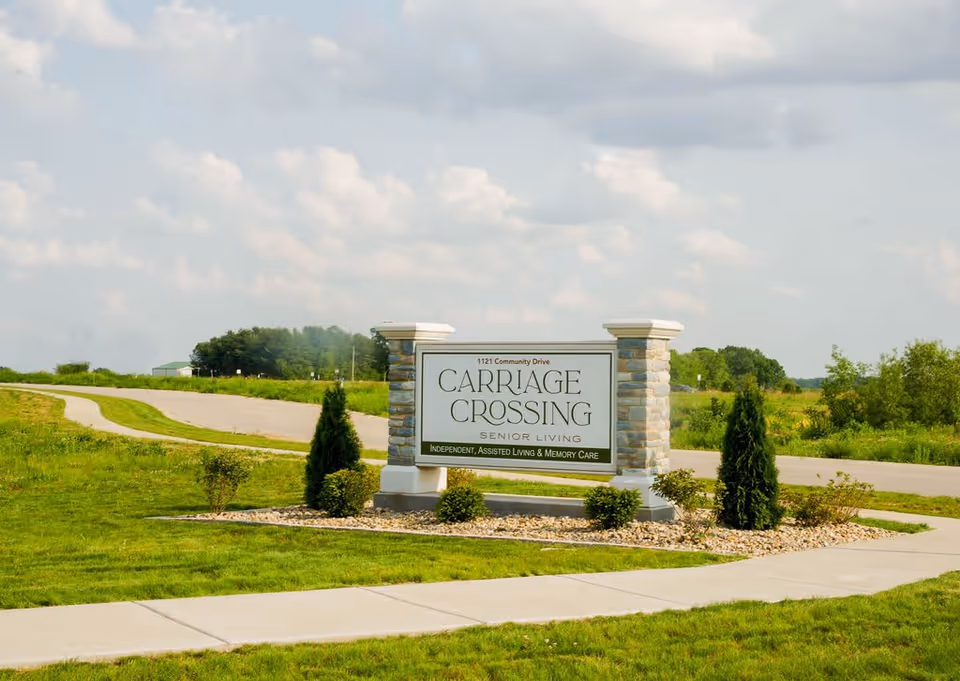 Stone entrance sign reading "Carriage Crossing Senior Living" on a grassy roadside at the facility entrance.