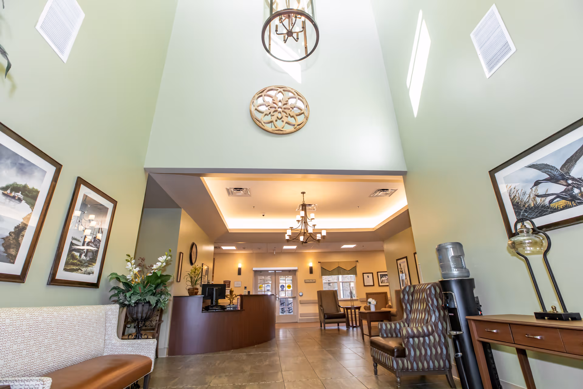 A spacious lobby of a senior living facility with a reception desk, seating, plants, and framed artwork under a vaulted ceiling.
