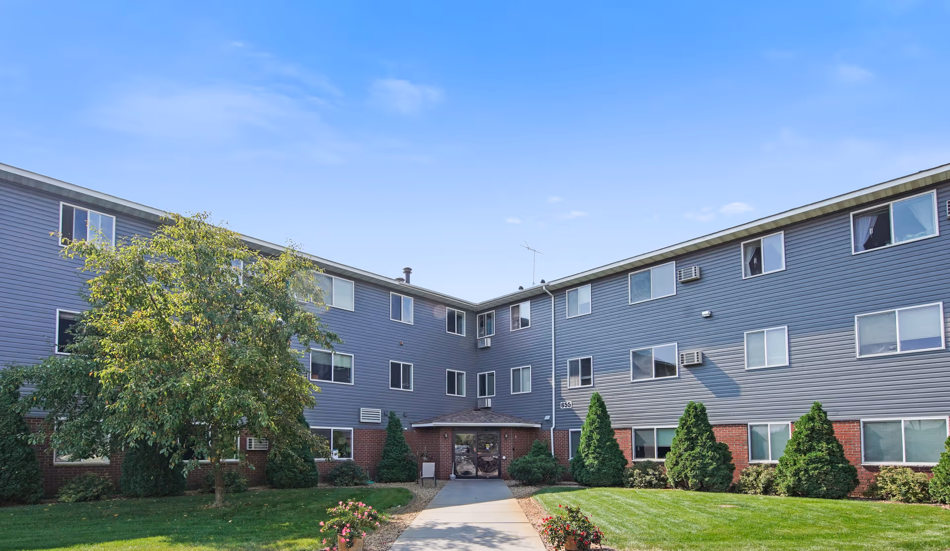 Exterior view of a three-story senior living facility building with gray siding and red brick accents. There is a paved walkway leading to the main entrance, surrounded by green grass, bushes, and a tree. The sky is clear and blue.