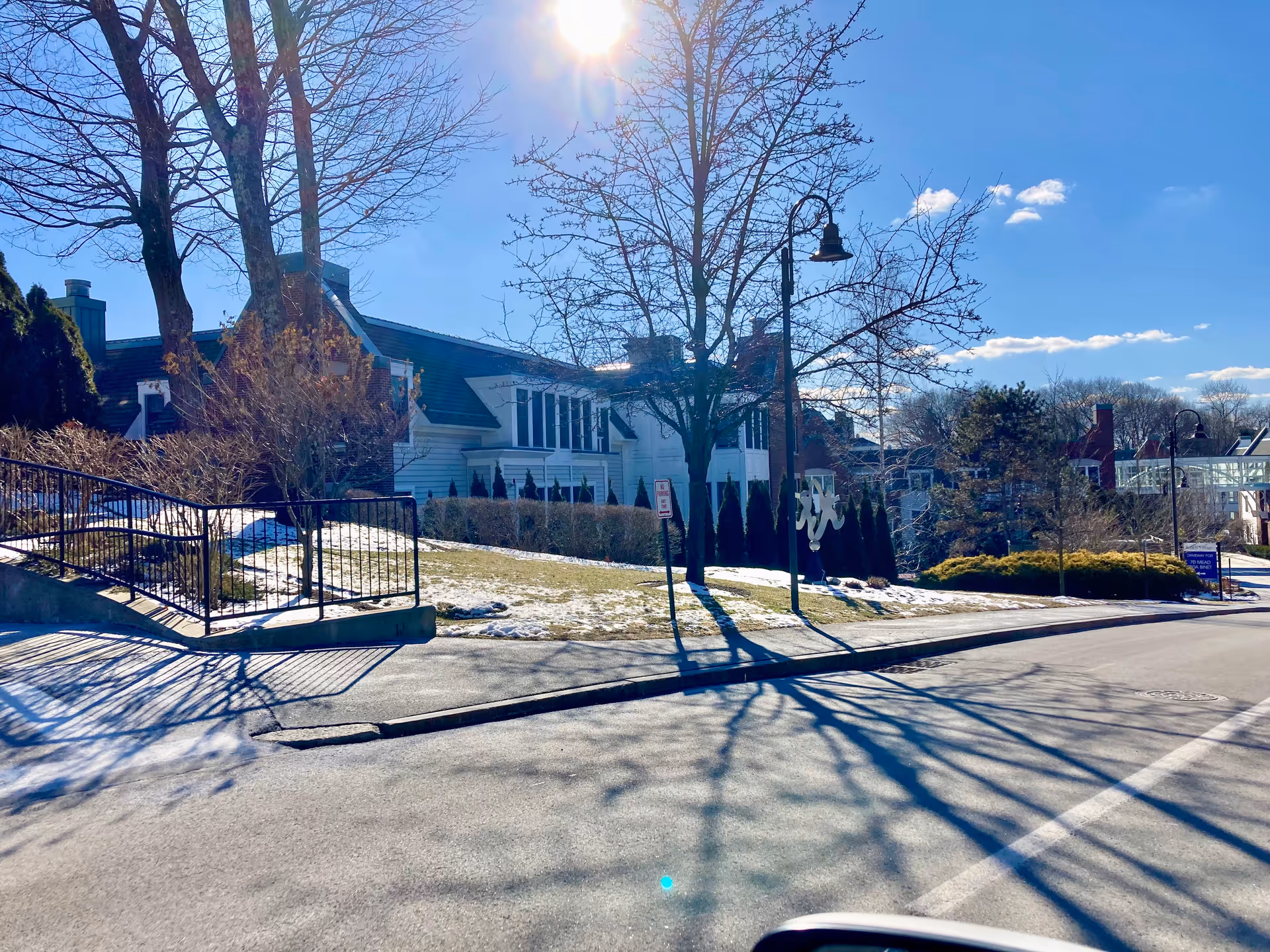 Exterior view of a senior living facility building with white siding and multiple windows. There are leafless trees casting shadows on the road and sidewalk, some patches of snow on the grass, and a clear blue sky with the sun shining brightly.