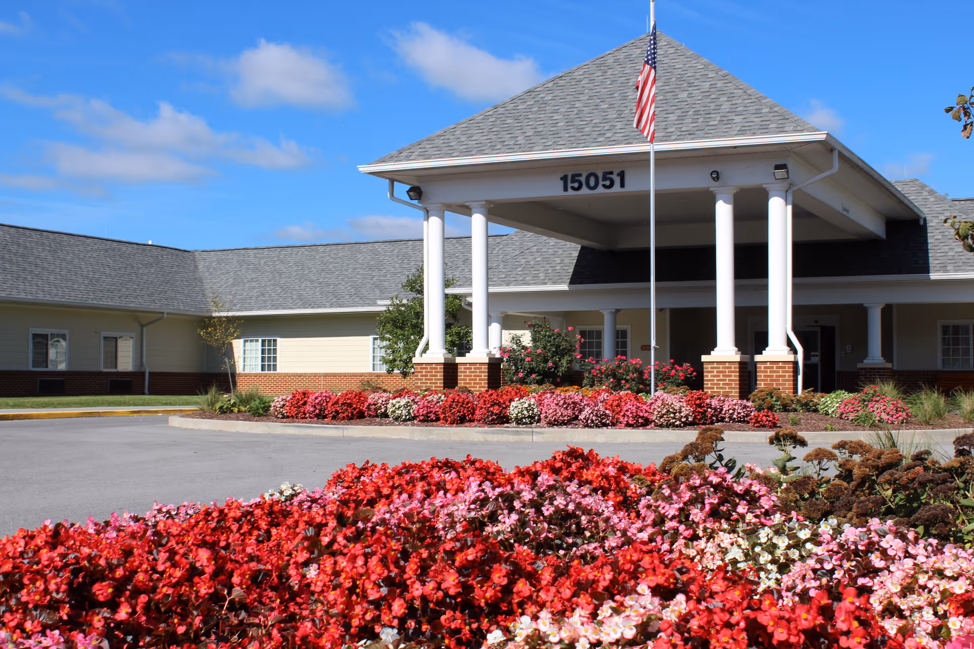 Front entrance of a single-story health and rehab building with a covered portico showing the number '15051', an American flag, and colorful flowerbeds.