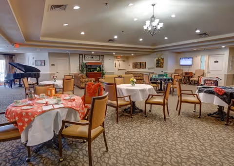 A spacious senior living facility common area with multiple round tables covered with tablecloths and set with cups and plates. There are several chairs around the tables, a grand piano in the corner, a fireplace with a TV mounted above it, and comfortable seating areas with armchairs and sofas. The room is well-lit with ceiling lights and a chandelier, and the carpet has a patterned design.