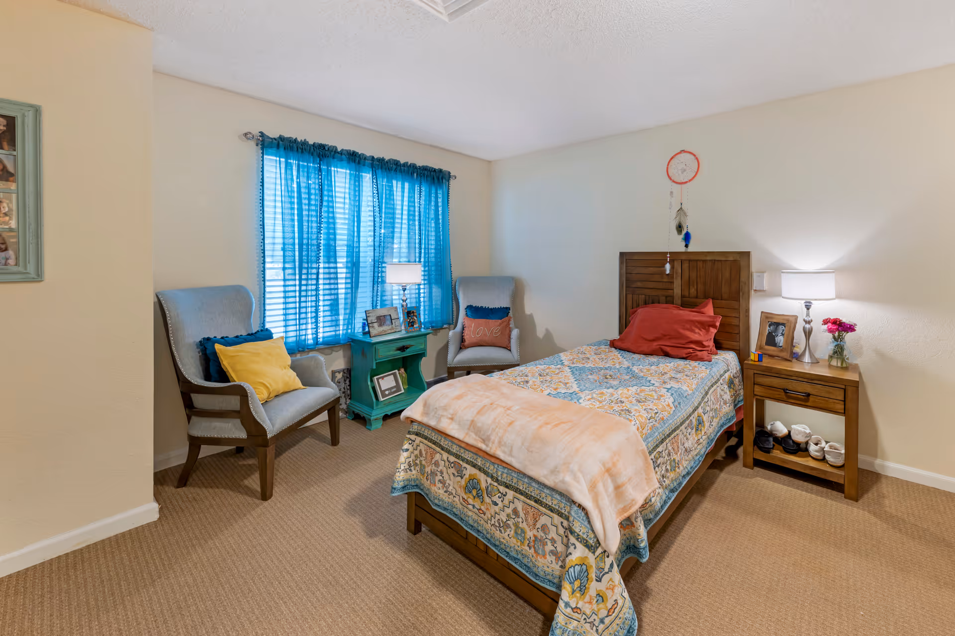 A cozy furnished bedroom with a single bed, wooden nightstand, two armchairs by a window with blue curtains, and decorative accents.