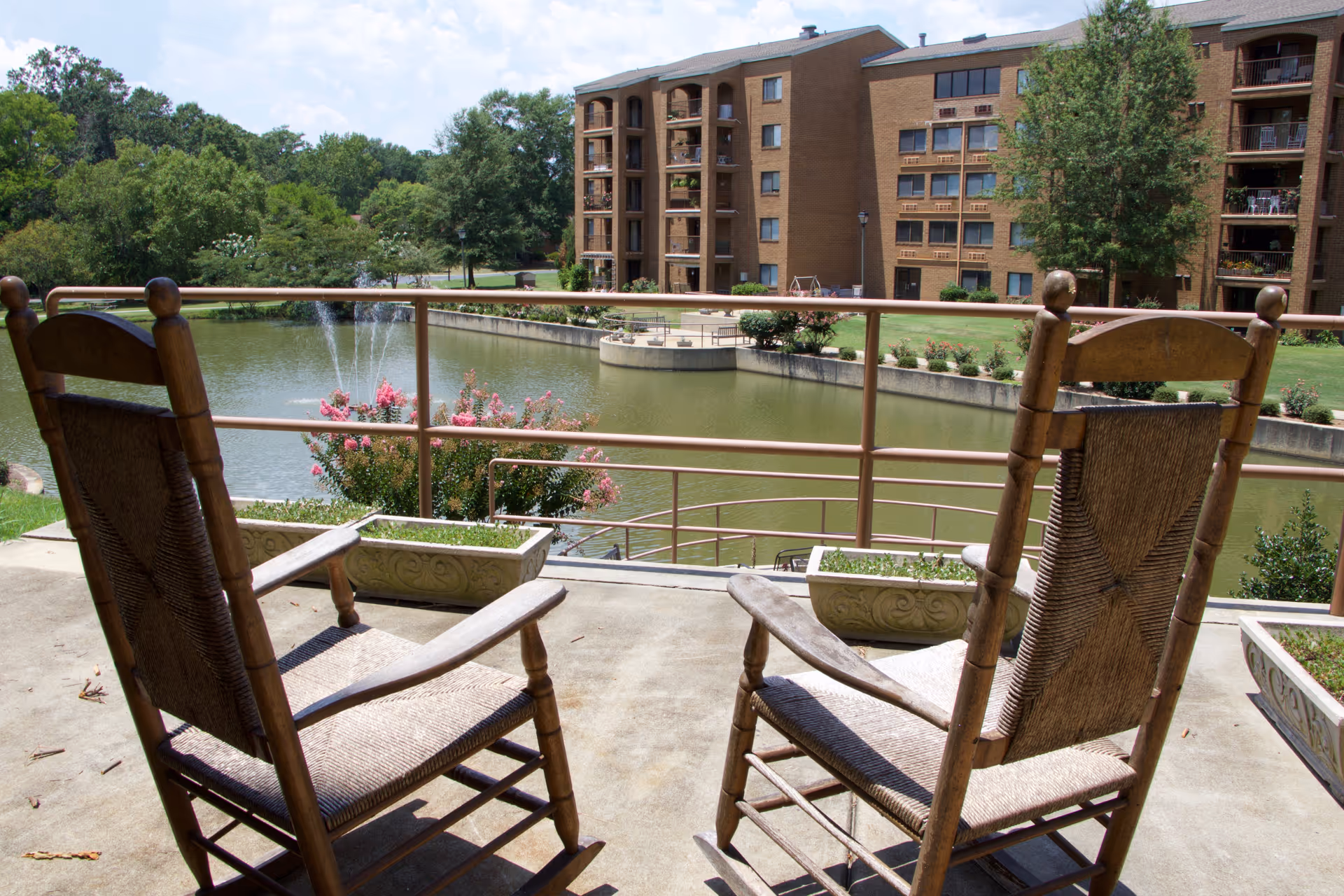 Two wooden rocking chairs on a concrete patio overlooking a pond with a water fountain, surrounded by greenery and a multi-story brick building in the background.