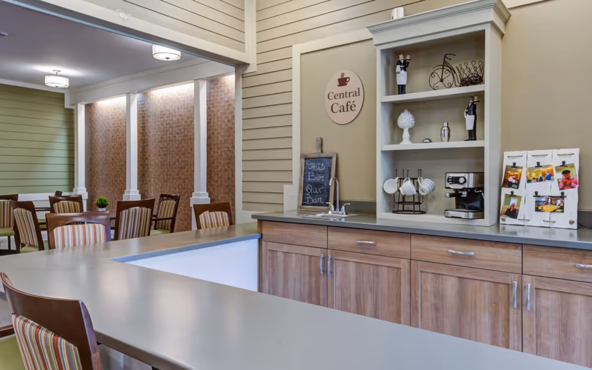 Interior view of a cozy café area in a senior living facility with a countertop, wooden cabinets, and shelves holding coffee mugs, a coffee machine, and decorative items. In the background, there are tables and chairs with striped upholstery arranged for dining or socializing, and a sign on the wall reads 'Central Café'.