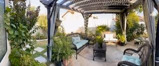 Outdoor patio area with a pergola overhead, surrounded by greenery and plants. The space includes cushioned chairs, a bench, and a small table, creating a comfortable seating area for relaxation.