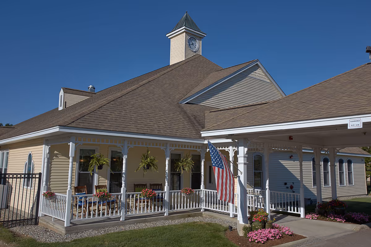 Exterior view of a senior living facility building with beige siding and a brown roof featuring a clock tower. The building has a covered porch with white railings, hanging plants, and rocking chairs. An American flag is displayed near the entrance, and there are flower beds with pink flowers along the walkway.