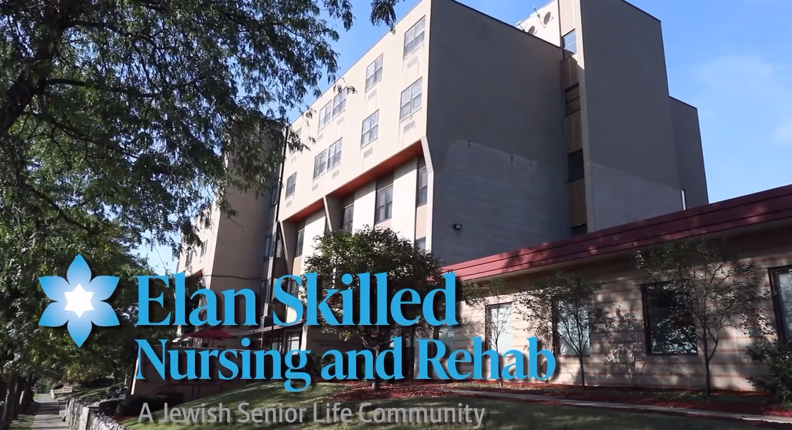 Exterior view of a multi-story building with beige and gray walls, surrounded by trees and greenery under a clear blue sky. The building has multiple windows and a red roof section near the entrance. Text overlay reads 'Elan Skilled Nursing and Rehab, A Jewish Senior Life Community.'