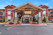 Front entrance and porte-cochère of The Welstone at Mission Crossing, a red multi-story senior living building with stone columns, landscaping, and an American flag.