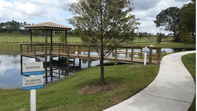 A wooden gazebo and dock over a pond with a paved walking path, a tree, and signs for a nature walk and no swimming.