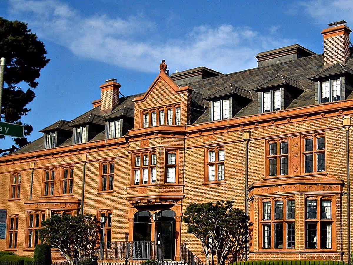Exterior view of a large, historic brick building with multiple windows and chimneys under a blue sky with some clouds. There are trimmed bushes in front of the building.