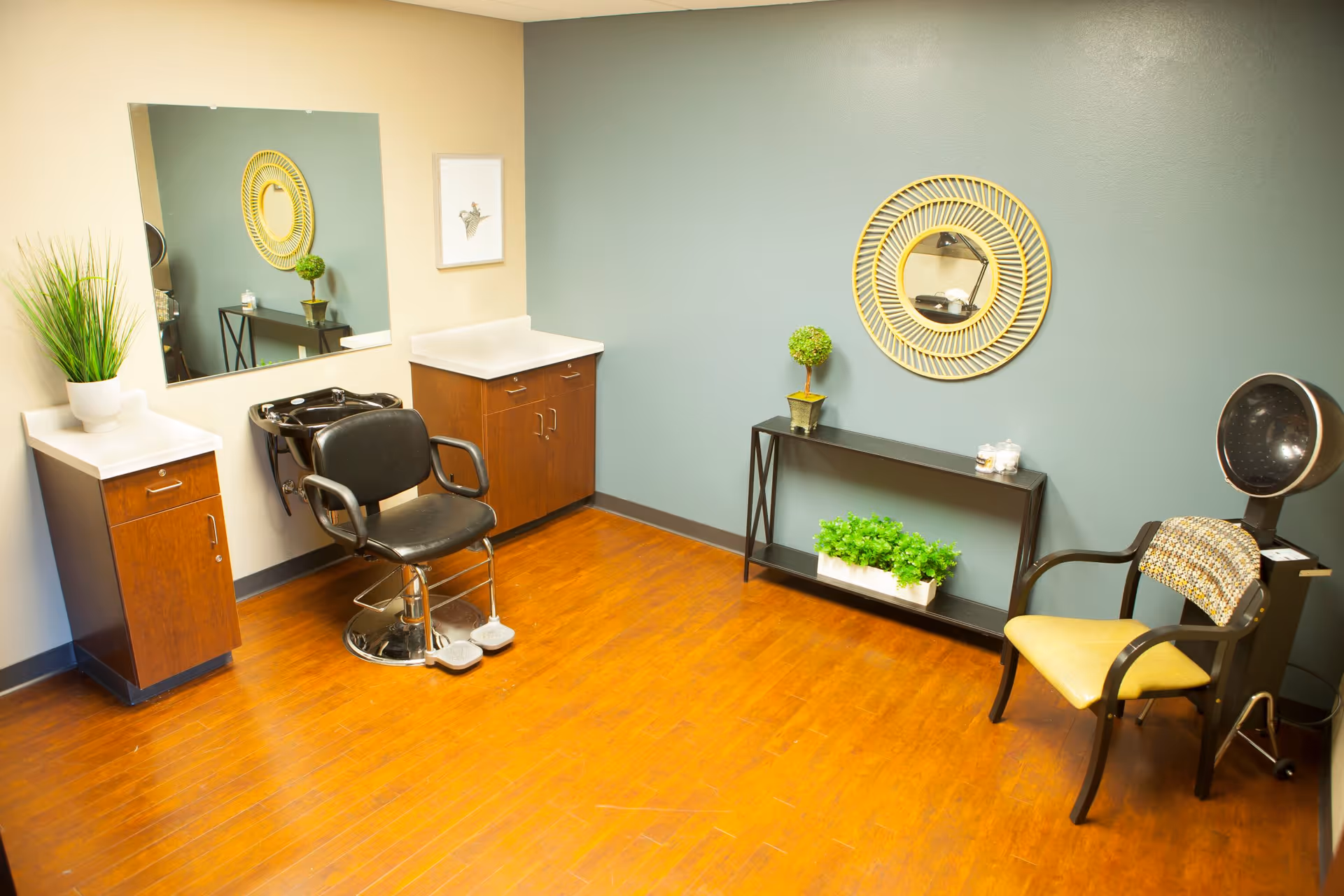 A bright and clean hair salon room with wooden flooring, a black salon chair in front of a black hair washing sink, two wooden cabinets with white countertops, a large wall mirror, a decorative round mirror with a yellow frame, a small black shelf with green plants, and a yellow cushioned chair next to a hair dryer.