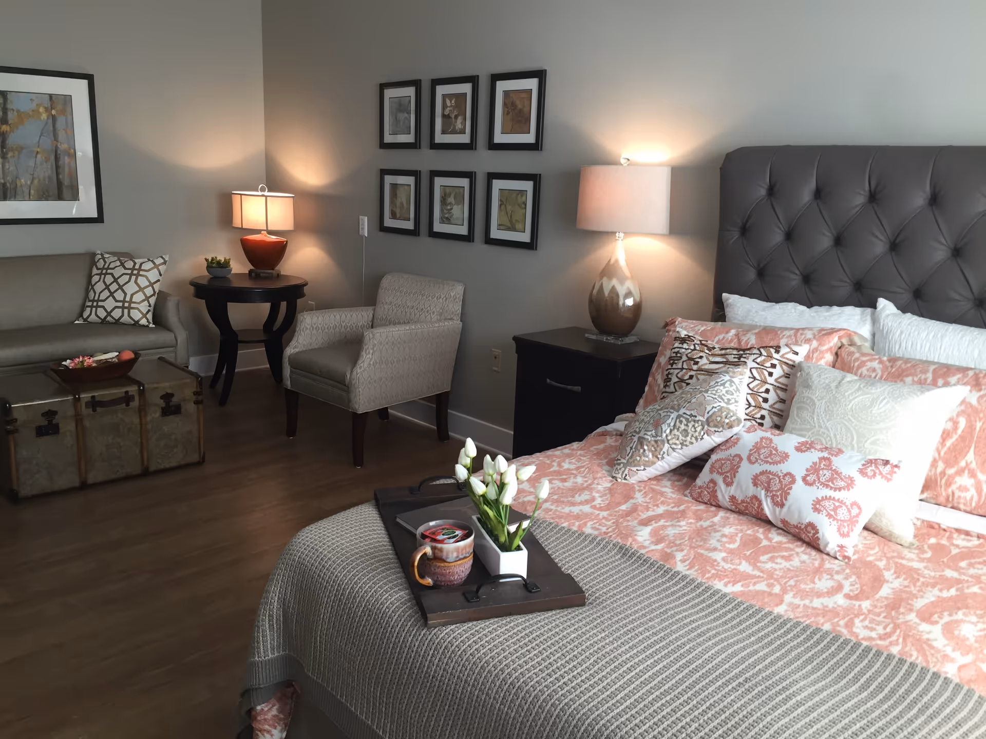 Stylish bedroom featuring a tufted headboard, patterned coral and gray bedding, bedside lamps, a seating area with chairs and a trunk, and hardwood floors.
