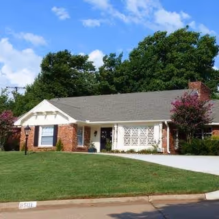 Single-story brick house with a gray shingled roof, white trim, and a well-maintained green lawn in front. There are trees and flowering bushes around the house under a blue sky with some clouds.