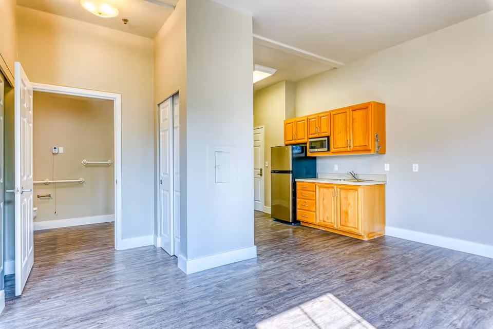 Interior view of a senior living facility unit showing a small kitchen area with wooden cabinets, a microwave, and a refrigerator. The room has light-colored walls and wood-style flooring. To the left, there is an open door leading to a bathroom equipped with grab bars for accessibility.