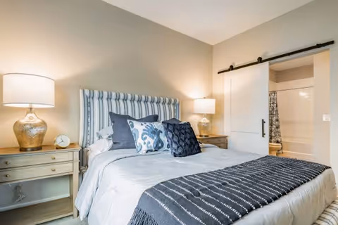 A neatly made bedroom with a striped upholstered headboard, bedside tables and lamps, decorative pillows, and a sliding barn door opening to a bathroom.