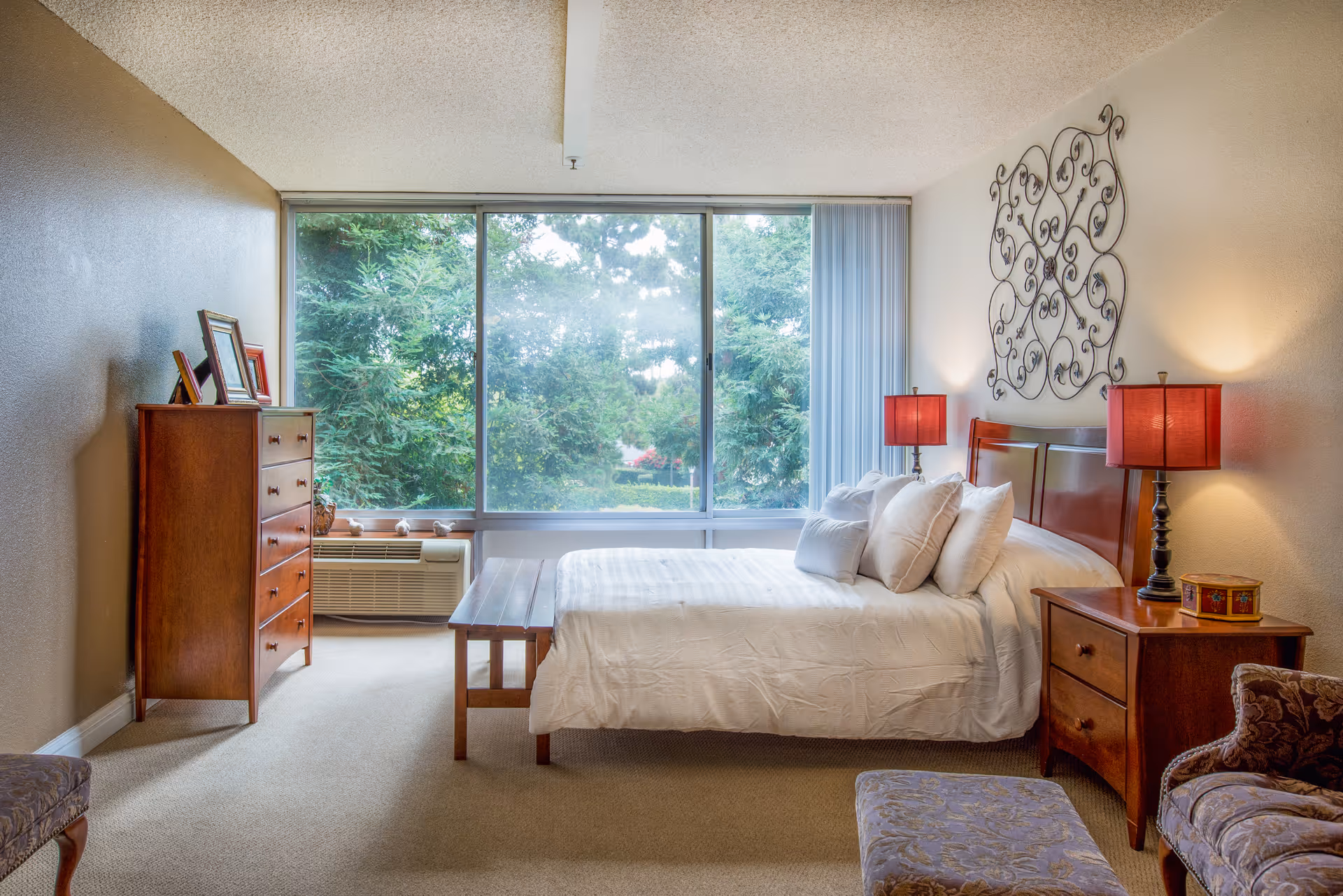 A cozy bedroom with a large window showing green trees outside. The room features a wooden bed with white bedding and multiple pillows, two red lamps on wooden nightstands, a wooden dresser with picture frames, and a patterned armchair with an ottoman.