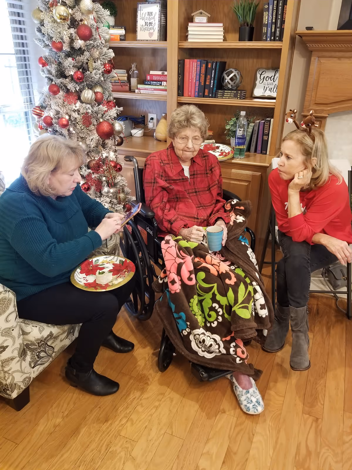 Three women gathered in a decorated common room with a Christmas tree, an elderly woman in a wheelchair holding a cup and a colorful blanket.