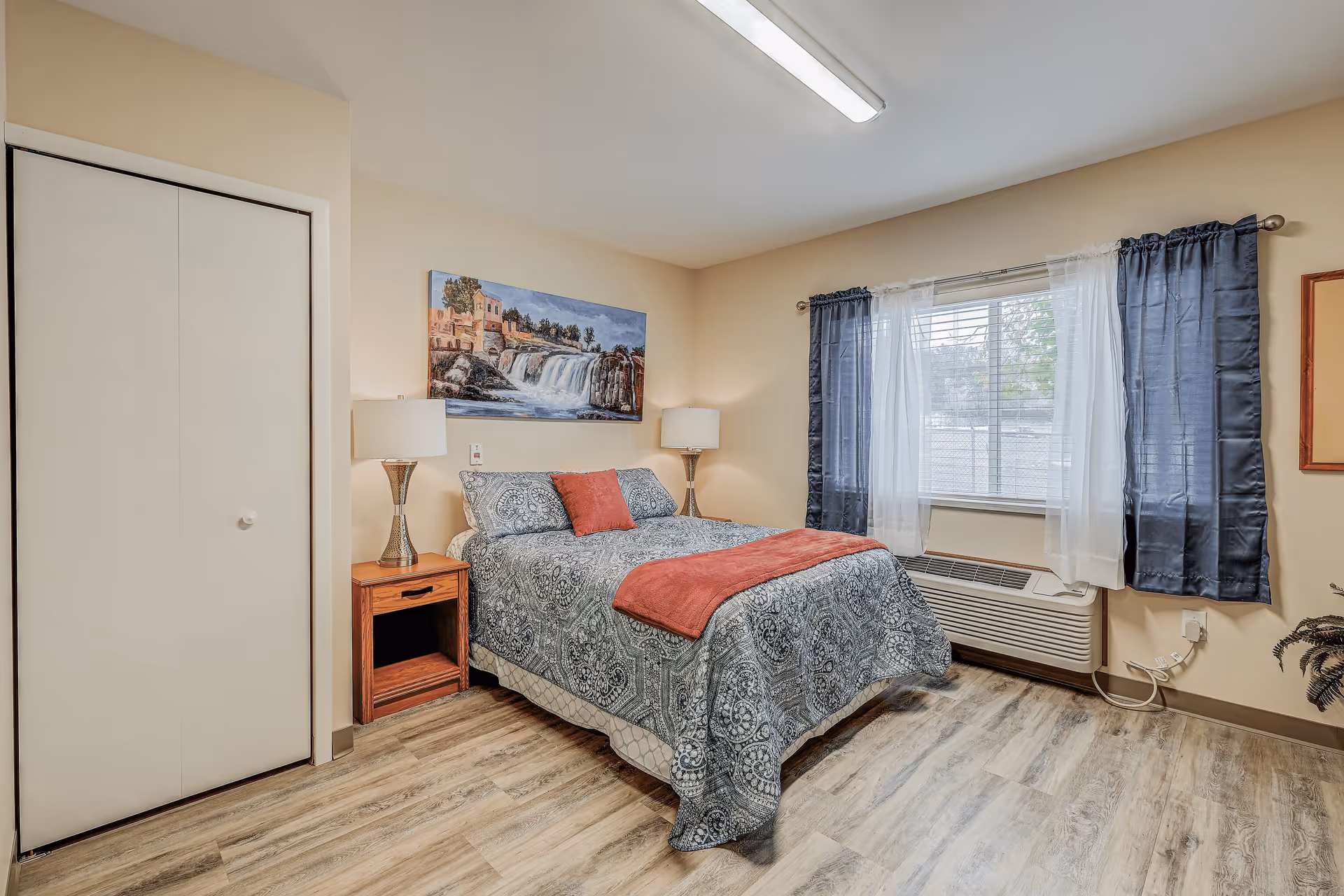 Clean, furnished bedroom featuring a bed with patterned bedding, two lamps, a nightstand, a window with curtains, and a closet.