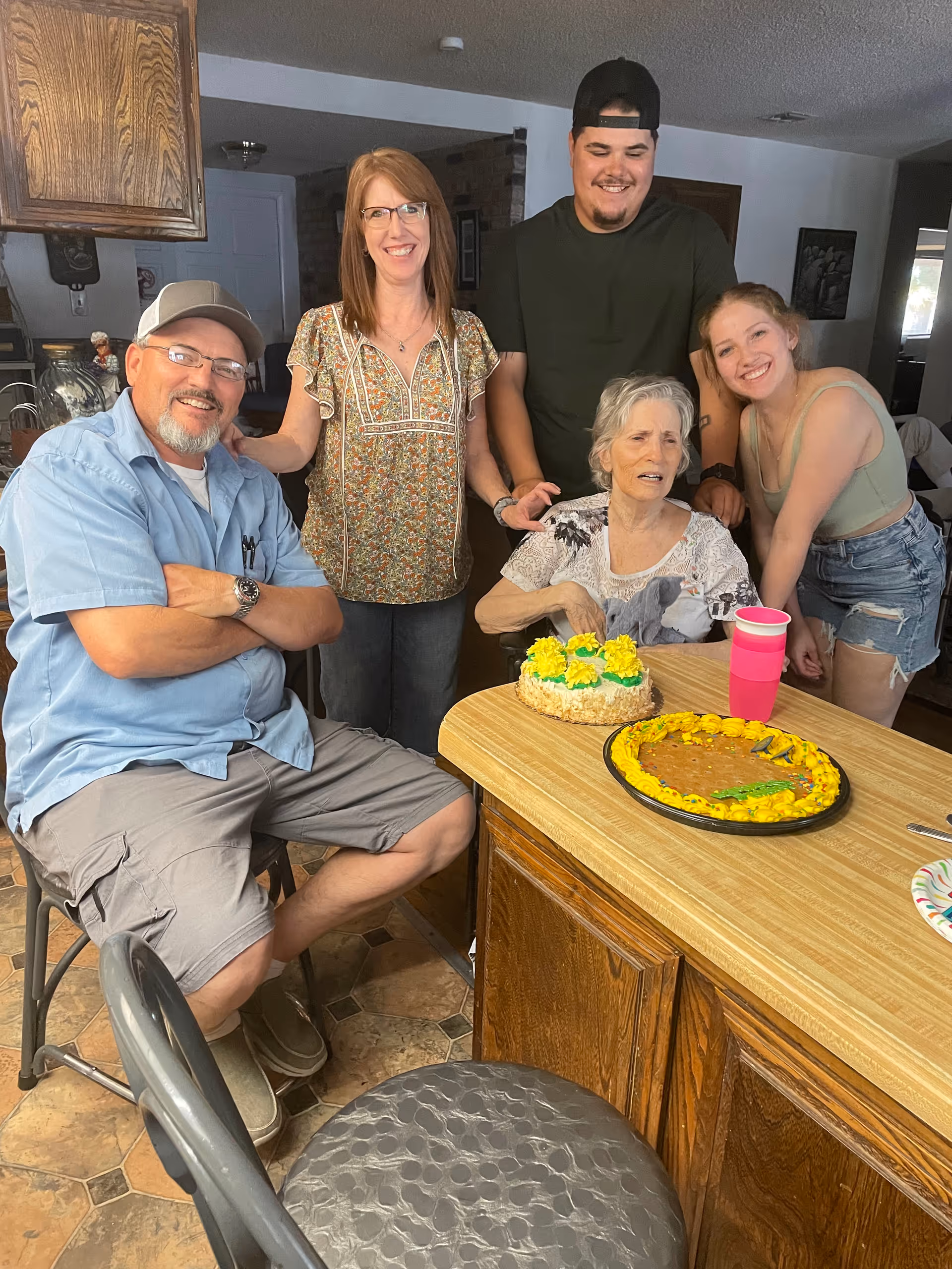 A group of five people gathered around a kitchen island with two decorated cakes and a pink cup on it. The group includes an elderly woman seated at the island, two women and two men standing and sitting around her, all smiling and appearing happy in a home kitchen setting.